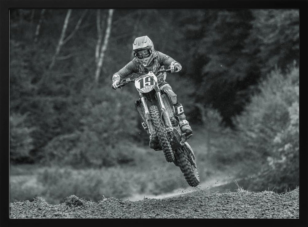 A dynamic black and white photograph of a motocross racer in mid-air, jumping over a dirt mound. The rider, wearing full protective gear and a helmet, is on a dirt bike with the number 19 plate. The background is a soft-focus forest, and the entire image is enclosed in a silver frame. Poster