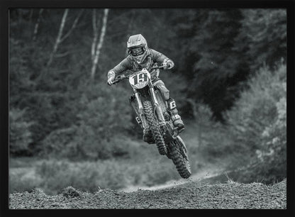 A dynamic black and white photograph of a motocross racer in mid-air, jumping over a dirt mound. The rider, wearing full protective gear and a helmet, is on a dirt bike with the number 19 plate. The background is a soft-focus forest, and the entire image is enclosed in a silver frame. Poster