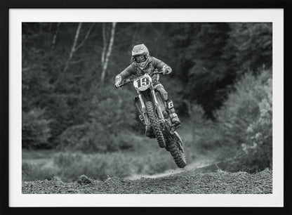 A dynamic black and white photograph of a motocross racer in mid-air, jumping over a dirt mound. The rider, wearing full protective gear and a helmet, is on a dirt bike with the number 19 plate. The background is a soft-focus forest, and the entire image is enclosed in a silver frame. Poster