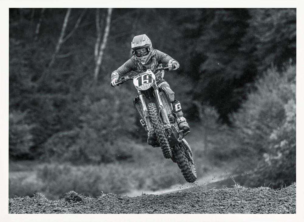 A dynamic black and white photograph of a motocross racer in mid-air, jumping over a dirt mound. The rider, wearing full protective gear and a helmet, is on a dirt bike with the number 19 plate. The background is a soft-focus forest, and the entire image is enclosed in a silver frame. Poster