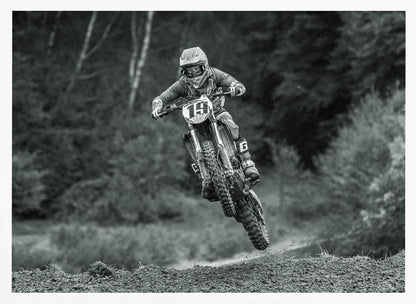 A dynamic black and white photograph of a motocross racer in mid-air, jumping over a dirt mound. The rider, wearing full protective gear and a helmet, is on a dirt bike with the number 19 plate. The background is a soft-focus forest, and the entire image is enclosed in a silver frame. Poster