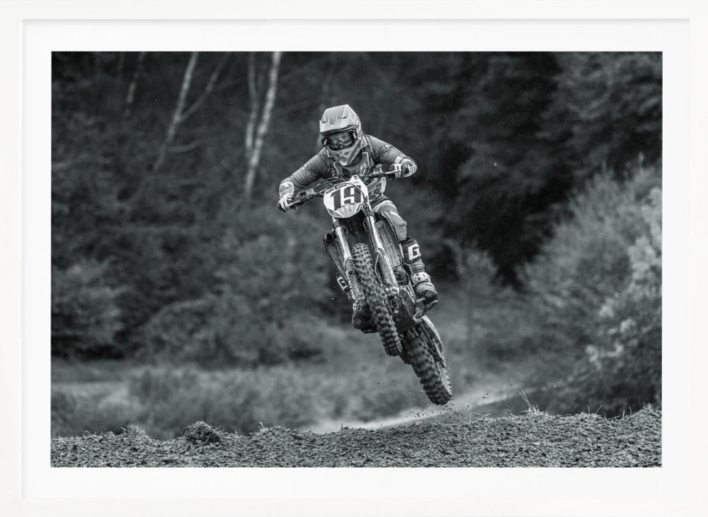 A dynamic black and white photograph of a motocross racer in mid-air, jumping over a dirt mound. The rider, wearing full protective gear and a helmet, is on a dirt bike with the number 19 plate. The background is a soft-focus forest, and the entire image is enclosed in a silver frame. Poster