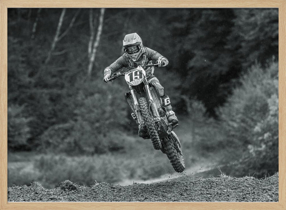 A dynamic black and white photograph of a motocross racer in mid-air, jumping over a dirt mound. The rider, wearing full protective gear and a helmet, is on a dirt bike with the number 19 plate. The background is a soft-focus forest, and the entire image is enclosed in a silver frame. Poster