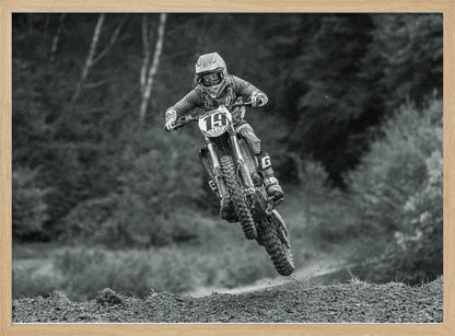 A dynamic black and white photograph of a motocross racer in mid-air, jumping over a dirt mound. The rider, wearing full protective gear and a helmet, is on a dirt bike with the number 19 plate. The background is a soft-focus forest, and the entire image is enclosed in a silver frame. Poster