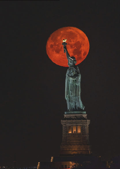 A striking night photograph of the Statue of Liberty perfectly silhouetted against a giant, vibrant red supermoon, which appears to be held aloft by the statue's torch against a deep black sky. Artwork