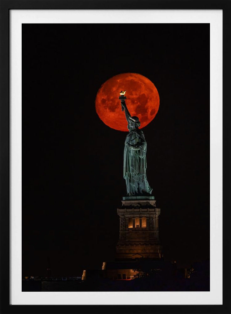 A striking night photograph of the Statue of Liberty perfectly silhouetted against a giant, vibrant red supermoon, which appears to be held aloft by the statue's torch against a deep black sky. Artwork