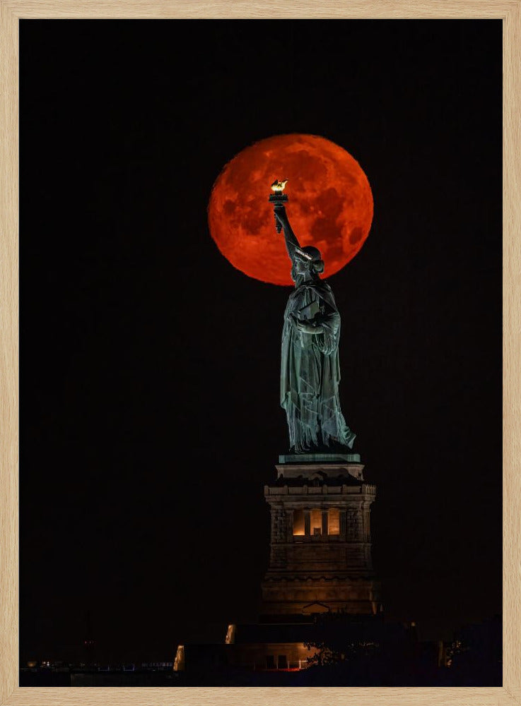 A striking night photograph of the Statue of Liberty perfectly silhouetted against a giant, vibrant red supermoon, which appears to be held aloft by the statue's torch against a deep black sky. Artwork