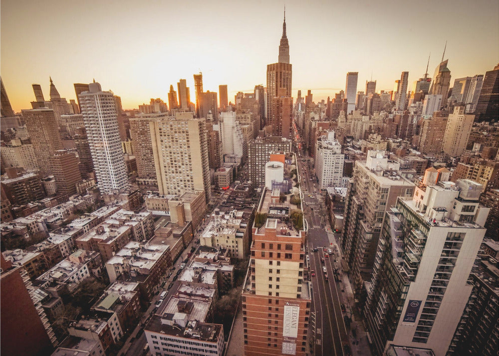 An aerial photograph of the New York City skyline at sunset, with the Empire State Building prominent against the glowing sky. The warm light of the setting sun bathes the dense urban landscape in a golden hue. The entire scene is enclosed in a silver, brushed metal frame. Print