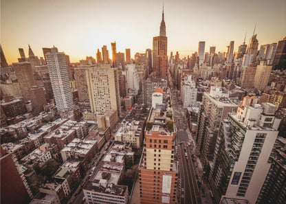 An aerial photograph of the New York City skyline at sunset, with the Empire State Building prominent against the glowing sky. The warm light of the setting sun bathes the dense urban landscape in a golden hue. The entire scene is enclosed in a silver, brushed metal frame. Print