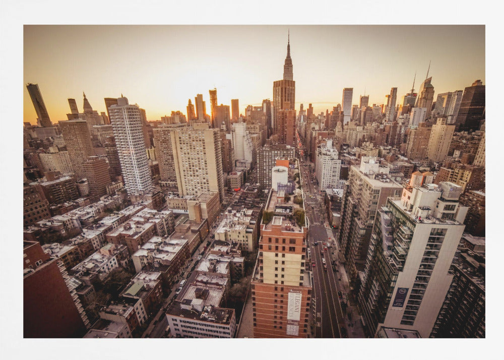 An aerial photograph of the New York City skyline at sunset, with the Empire State Building prominent against the glowing sky. The warm light of the setting sun bathes the dense urban landscape in a golden hue. The entire scene is enclosed in a silver, brushed metal frame. Print