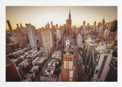 An aerial photograph of the New York City skyline at sunset, with the Empire State Building prominent against the glowing sky. The warm light of the setting sun bathes the dense urban landscape in a golden hue. The entire scene is enclosed in a silver, brushed metal frame. Print