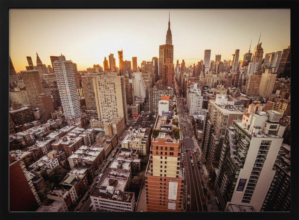 An aerial photograph of the New York City skyline at sunset, with the Empire State Building prominent against the glowing sky. The warm light of the setting sun bathes the dense urban landscape in a golden hue. The entire scene is enclosed in a silver, brushed metal frame. Print