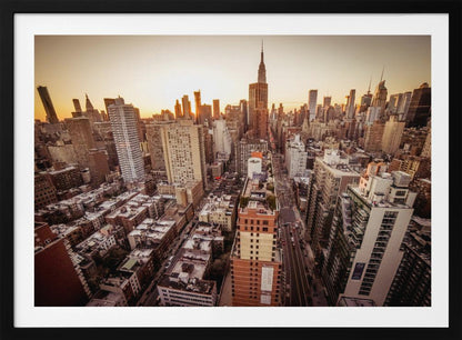 An aerial photograph of the New York City skyline at sunset, with the Empire State Building prominent against the glowing sky. The warm light of the setting sun bathes the dense urban landscape in a golden hue. The entire scene is enclosed in a silver, brushed metal frame. Print