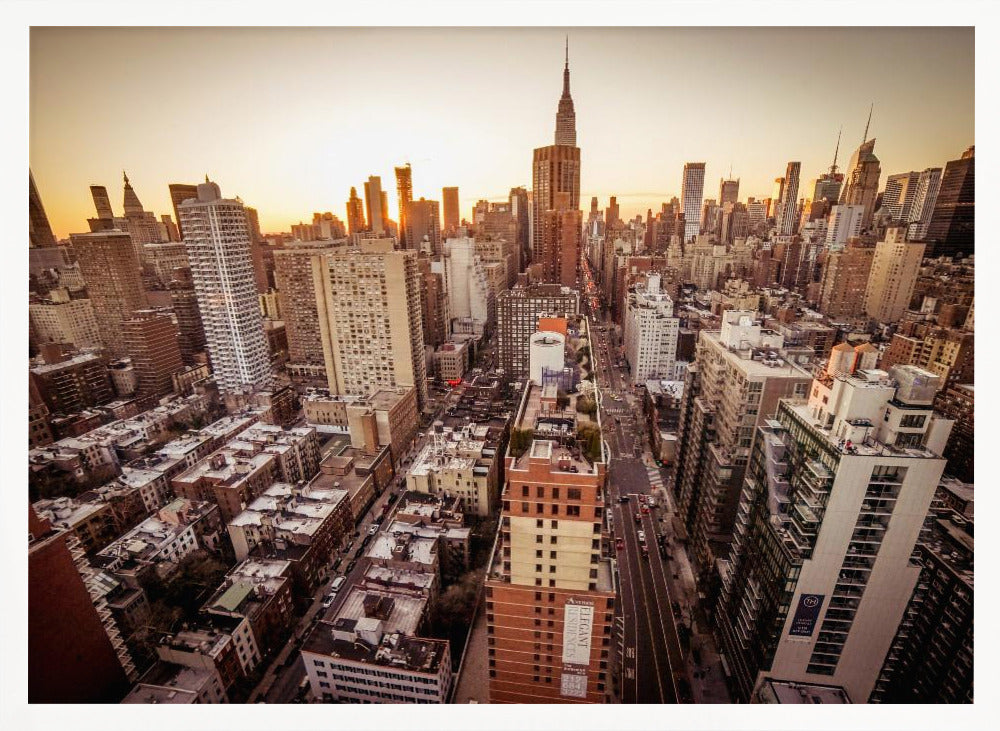 An aerial photograph of the New York City skyline at sunset, with the Empire State Building prominent against the glowing sky. The warm light of the setting sun bathes the dense urban landscape in a golden hue. The entire scene is enclosed in a silver, brushed metal frame. Print
