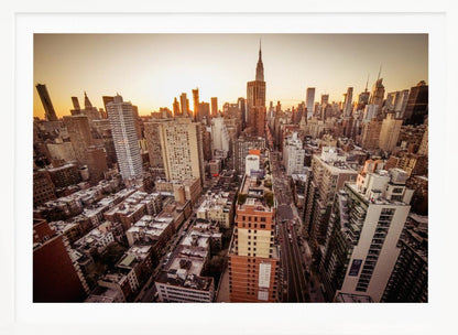 An aerial photograph of the New York City skyline at sunset, with the Empire State Building prominent against the glowing sky. The warm light of the setting sun bathes the dense urban landscape in a golden hue. The entire scene is enclosed in a silver, brushed metal frame. Print