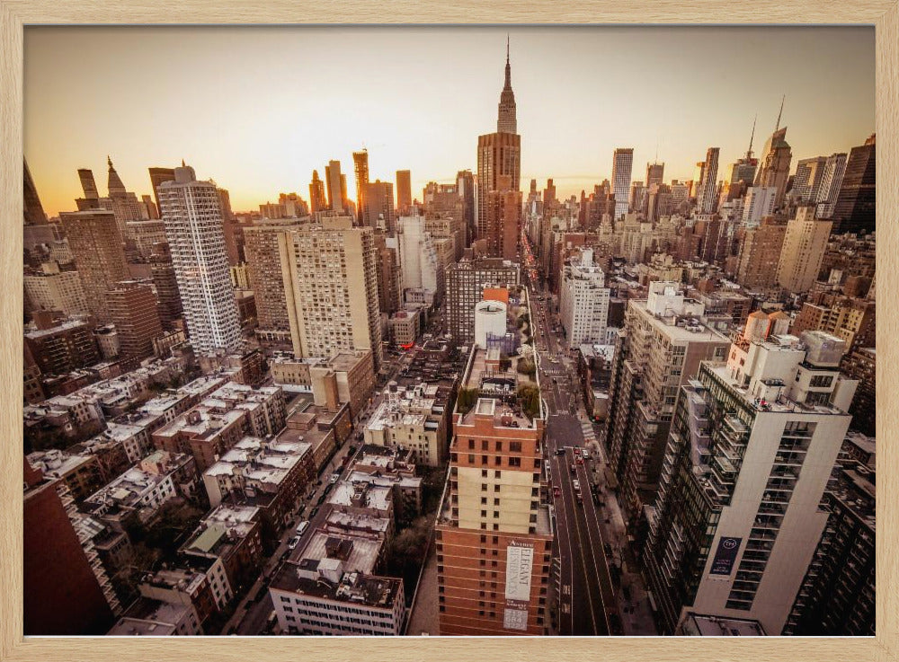 An aerial photograph of the New York City skyline at sunset, with the Empire State Building prominent against the glowing sky. The warm light of the setting sun bathes the dense urban landscape in a golden hue. The entire scene is enclosed in a silver, brushed metal frame. Print