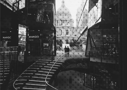 A framed, black and white photograph capturing a layered urban scene. The view, seen through a window with reflections of a curved, lit staircase, shows two people walking towards a large domed cathedral framed by modern glass buildings. Wall Art