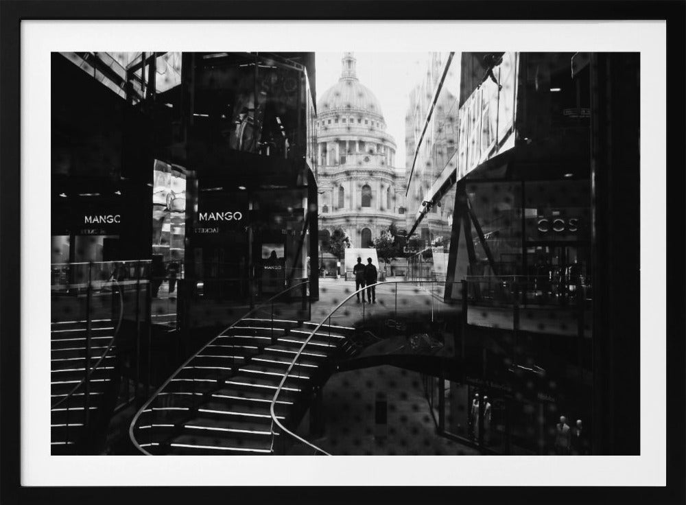 A framed, black and white photograph capturing a layered urban scene. The view, seen through a window with reflections of a curved, lit staircase, shows two people walking towards a large domed cathedral framed by modern glass buildings. Wall Art