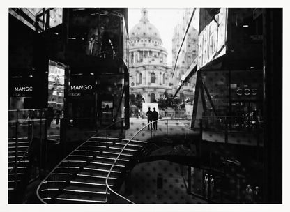 A framed, black and white photograph capturing a layered urban scene. The view, seen through a window with reflections of a curved, lit staircase, shows two people walking towards a large domed cathedral framed by modern glass buildings. Wall Art