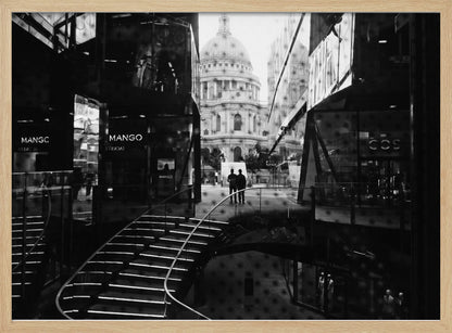 A framed, black and white photograph capturing a layered urban scene. The view, seen through a window with reflections of a curved, lit staircase, shows two people walking towards a large domed cathedral framed by modern glass buildings. Wall Art