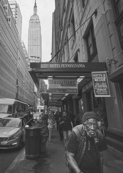 A powerful black and white vertical photograph capturing a New York City street scene. An elderly Black man with a weary expression is in the foreground, walking along a crowded sidewalk. In the background, the Empire State Building towers over the cityscape, framed between buildings, with a sign for the Hotel Pennsylvania visible. Artwork