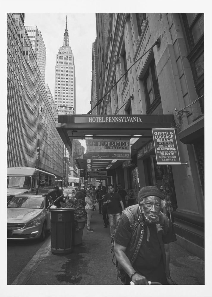 A powerful black and white vertical photograph capturing a New York City street scene. An elderly Black man with a weary expression is in the foreground, walking along a crowded sidewalk. In the background, the Empire State Building towers over the cityscape, framed between buildings, with a sign for the Hotel Pennsylvania visible. Artwork