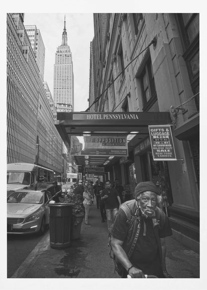 A powerful black and white vertical photograph capturing a New York City street scene. An elderly Black man with a weary expression is in the foreground, walking along a crowded sidewalk. In the background, the Empire State Building towers over the cityscape, framed between buildings, with a sign for the Hotel Pennsylvania visible. Artwork