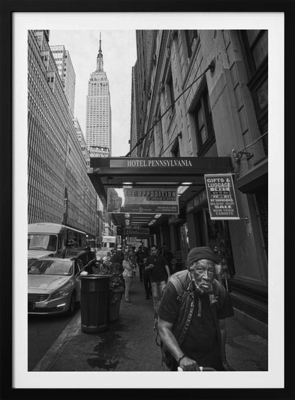 A powerful black and white vertical photograph capturing a New York City street scene. An elderly Black man with a weary expression is in the foreground, walking along a crowded sidewalk. In the background, the Empire State Building towers over the cityscape, framed between buildings, with a sign for the Hotel Pennsylvania visible. Artwork
