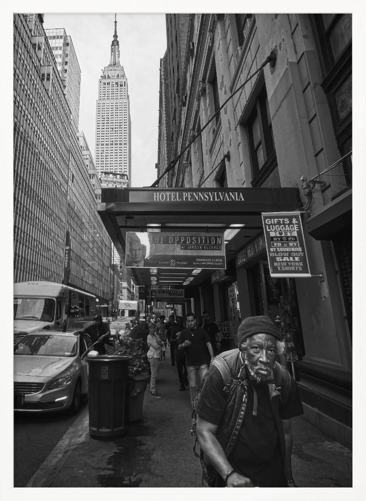 A powerful black and white vertical photograph capturing a New York City street scene. An elderly Black man with a weary expression is in the foreground, walking along a crowded sidewalk. In the background, the Empire State Building towers over the cityscape, framed between buildings, with a sign for the Hotel Pennsylvania visible. Artwork