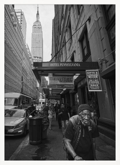 A powerful black and white vertical photograph capturing a New York City street scene. An elderly Black man with a weary expression is in the foreground, walking along a crowded sidewalk. In the background, the Empire State Building towers over the cityscape, framed between buildings, with a sign for the Hotel Pennsylvania visible. Artwork