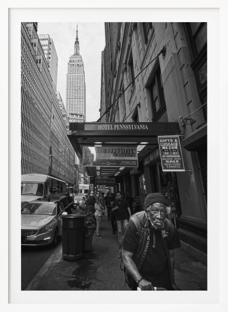 A powerful black and white vertical photograph capturing a New York City street scene. An elderly Black man with a weary expression is in the foreground, walking along a crowded sidewalk. In the background, the Empire State Building towers over the cityscape, framed between buildings, with a sign for the Hotel Pennsylvania visible. Artwork