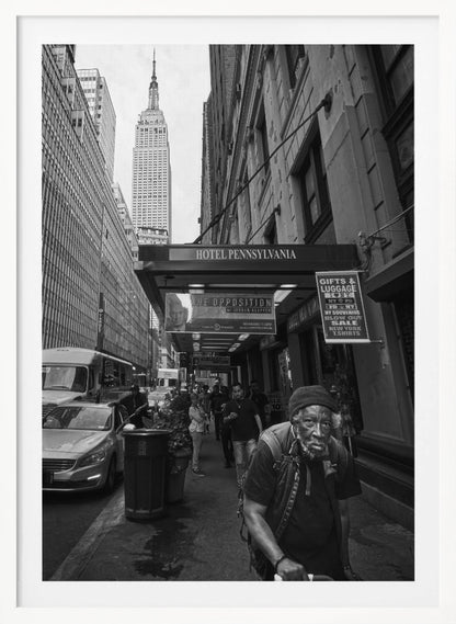 A powerful black and white vertical photograph capturing a New York City street scene. An elderly Black man with a weary expression is in the foreground, walking along a crowded sidewalk. In the background, the Empire State Building towers over the cityscape, framed between buildings, with a sign for the Hotel Pennsylvania visible. Artwork