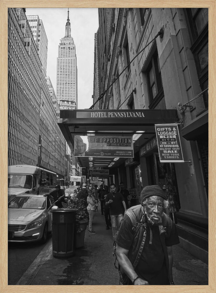 A powerful black and white vertical photograph capturing a New York City street scene. An elderly Black man with a weary expression is in the foreground, walking along a crowded sidewalk. In the background, the Empire State Building towers over the cityscape, framed between buildings, with a sign for the Hotel Pennsylvania visible. Artwork