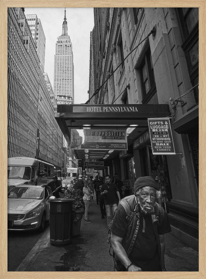 A powerful black and white vertical photograph capturing a New York City street scene. An elderly Black man with a weary expression is in the foreground, walking along a crowded sidewalk. In the background, the Empire State Building towers over the cityscape, framed between buildings, with a sign for the Hotel Pennsylvania visible. Artwork
