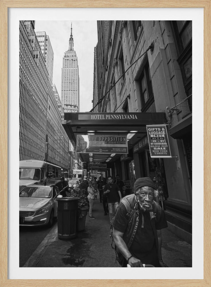 A powerful black and white vertical photograph capturing a New York City street scene. An elderly Black man with a weary expression is in the foreground, walking along a crowded sidewalk. In the background, the Empire State Building towers over the cityscape, framed between buildings, with a sign for the Hotel Pennsylvania visible. Artwork