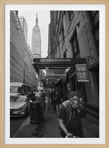 A powerful black and white vertical photograph capturing a New York City street scene. An elderly Black man with a weary expression is in the foreground, walking along a crowded sidewalk. In the background, the Empire State Building towers over the cityscape, framed between buildings, with a sign for the Hotel Pennsylvania visible. Artwork