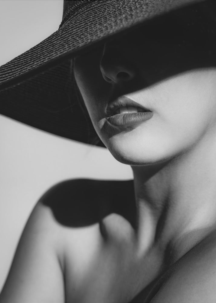 A dramatic black and white close-up photograph of a woman wearing a large, dark, wide-brimmed hat. The hat casts a deep shadow over the upper half of her face, obscuring her eyes and creating a sense of mystery. Only her nose, full lips, chin, neck, and shoulder are visible, highlighted by a strong light source. Decor