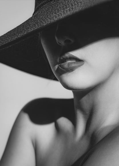 A dramatic black and white close-up photograph of a woman wearing a large, dark, wide-brimmed hat. The hat casts a deep shadow over the upper half of her face, obscuring her eyes and creating a sense of mystery. Only her nose, full lips, chin, neck, and shoulder are visible, highlighted by a strong light source. Decor