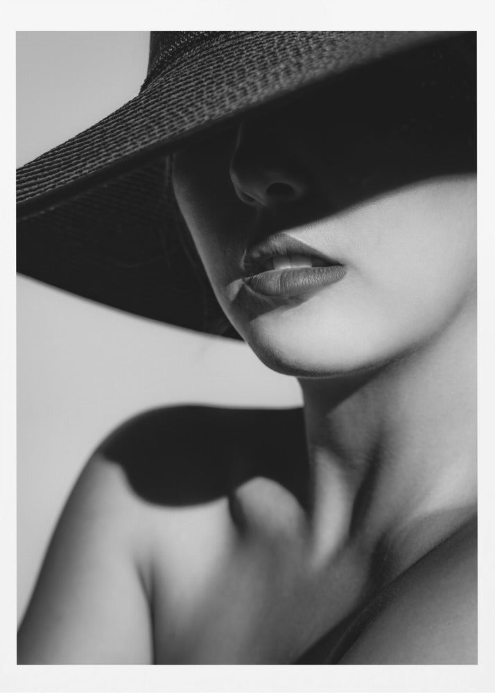 A dramatic black and white close-up photograph of a woman wearing a large, dark, wide-brimmed hat. The hat casts a deep shadow over the upper half of her face, obscuring her eyes and creating a sense of mystery. Only her nose, full lips, chin, neck, and shoulder are visible, highlighted by a strong light source. Decor