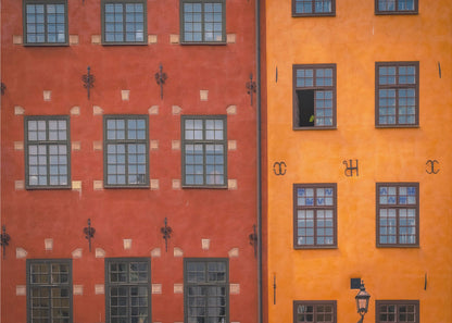A framed close-up photograph of two adjacent, colorful building facades. The building on the left is a deep, rustic red, while the one on the right is a vibrant orange. Both feature numerous multi-paned windows with dark frames. Artwork