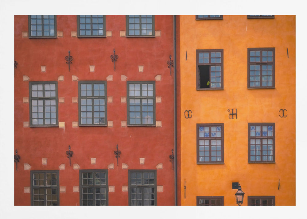 A framed close-up photograph of two adjacent, colorful building facades. The building on the left is a deep, rustic red, while the one on the right is a vibrant orange. Both feature numerous multi-paned windows with dark frames. Artwork