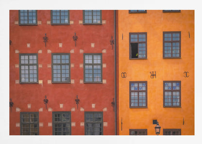 A framed close-up photograph of two adjacent, colorful building facades. The building on the left is a deep, rustic red, while the one on the right is a vibrant orange. Both feature numerous multi-paned windows with dark frames. Artwork
