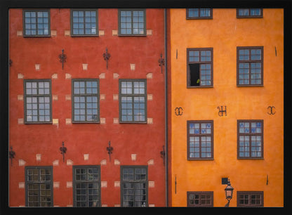 A framed close-up photograph of two adjacent, colorful building facades. The building on the left is a deep, rustic red, while the one on the right is a vibrant orange. Both feature numerous multi-paned windows with dark frames. Artwork