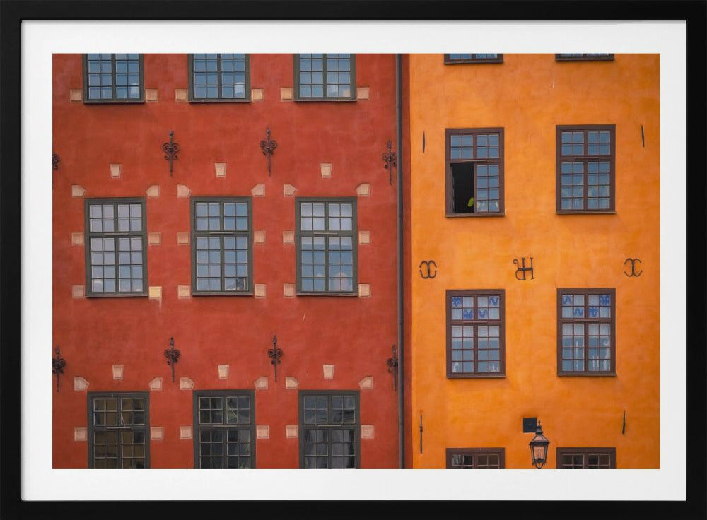 A framed close-up photograph of two adjacent, colorful building facades. The building on the left is a deep, rustic red, while the one on the right is a vibrant orange. Both feature numerous multi-paned windows with dark frames. Artwork