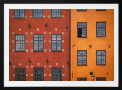 A framed close-up photograph of two adjacent, colorful building facades. The building on the left is a deep, rustic red, while the one on the right is a vibrant orange. Both feature numerous multi-paned windows with dark frames. Artwork