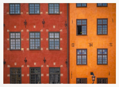 A framed close-up photograph of two adjacent, colorful building facades. The building on the left is a deep, rustic red, while the one on the right is a vibrant orange. Both feature numerous multi-paned windows with dark frames. Artwork