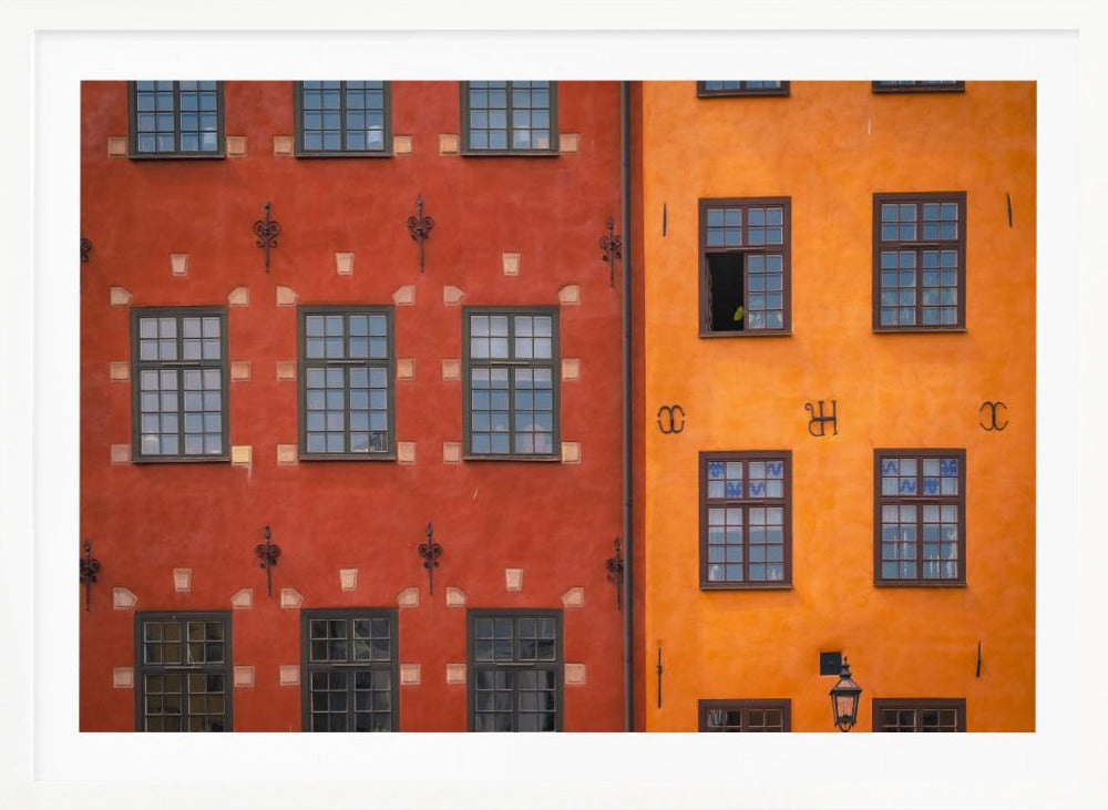 A framed close-up photograph of two adjacent, colorful building facades. The building on the left is a deep, rustic red, while the one on the right is a vibrant orange. Both feature numerous multi-paned windows with dark frames. Artwork