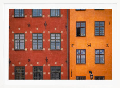 A framed close-up photograph of two adjacent, colorful building facades. The building on the left is a deep, rustic red, while the one on the right is a vibrant orange. Both feature numerous multi-paned windows with dark frames. Artwork