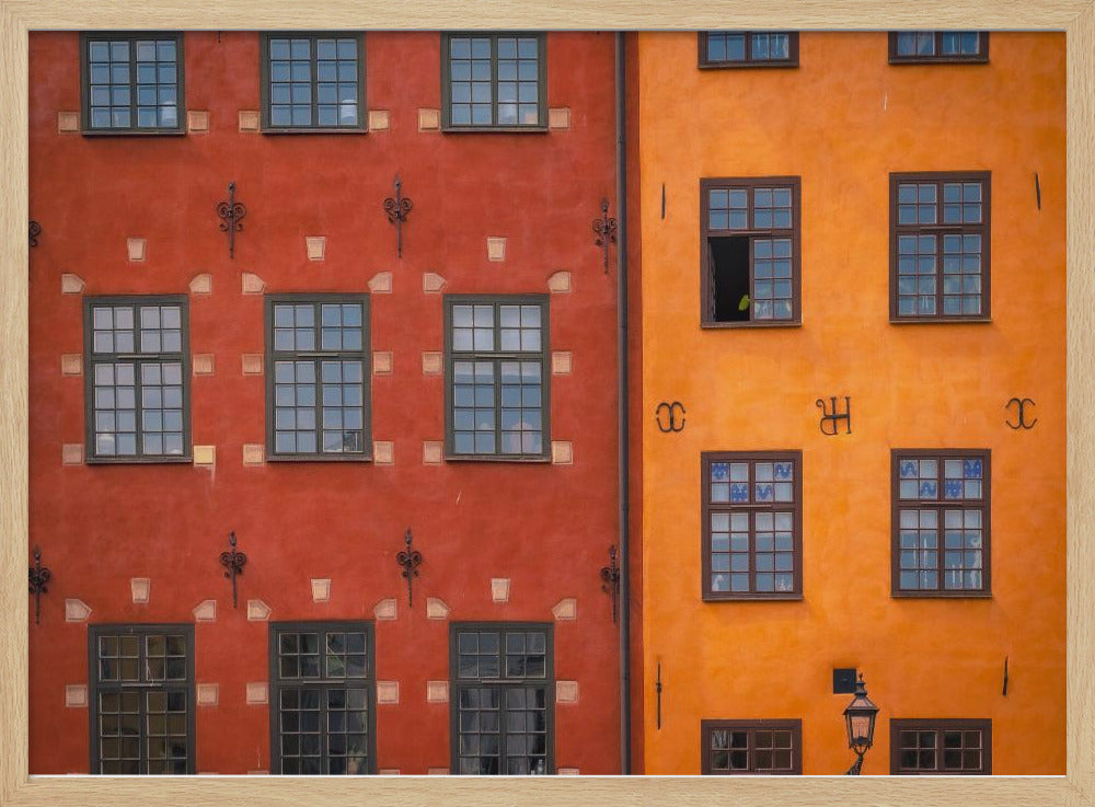 A framed close-up photograph of two adjacent, colorful building facades. The building on the left is a deep, rustic red, while the one on the right is a vibrant orange. Both feature numerous multi-paned windows with dark frames. Artwork