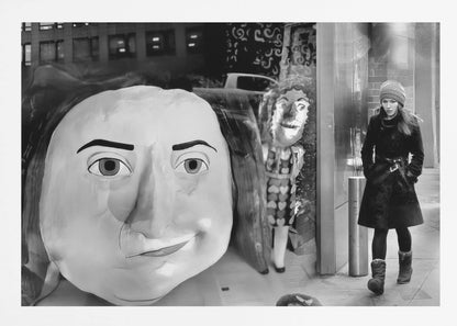 A surreal black and white photograph with a silver frame, featuring a giant, cartoonish head of a historical figure reflected in a window, juxtaposed with a young woman in a winter coat and beanie walking down a city sidewalk. Wall Art